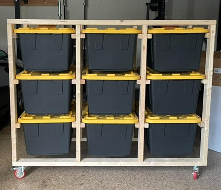 A cleanly organized garage featuring one wooden bin storage unit that is holding 9 black and yellow storage bins. The unit is 3 wide x 3 high and sits on top of casters.