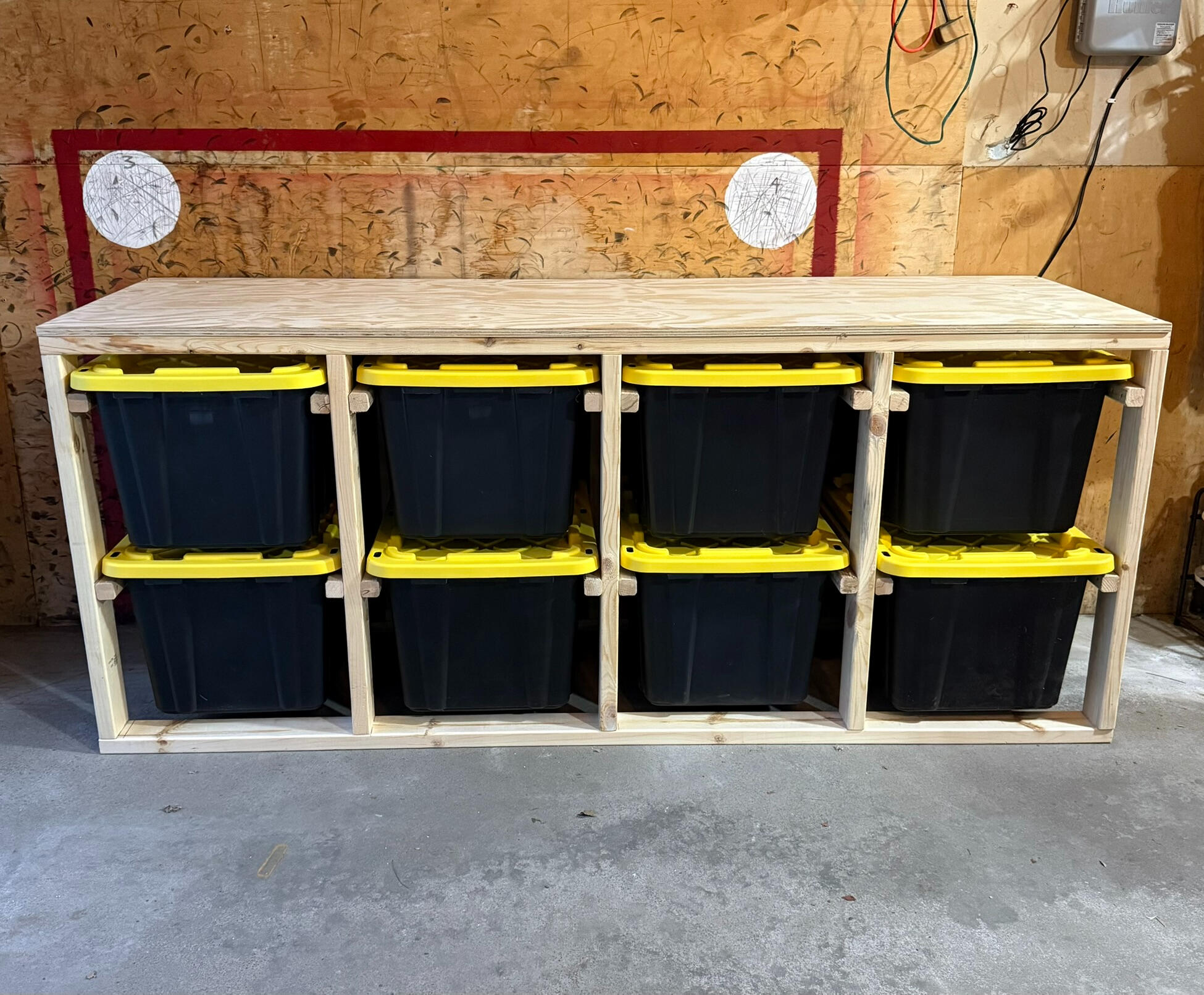 A cleanly organized garage featuring one wooden bin storage unit that is holding 9 black and yellow storage bins. The unit is 3 wide x 3 high and sits on top of casters.